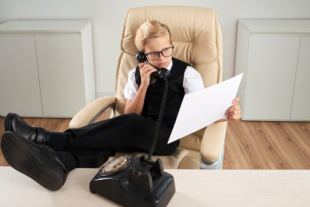 Menino caucasiano, sentado no escritório, na cadeira executiva, com os pés na mesa e falando no telefone