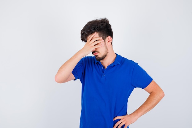 Menino cobrindo parte do rosto com a mão, mantendo a outra mão na cintura em uma camiseta azul e parecendo irritado. vista frontal.