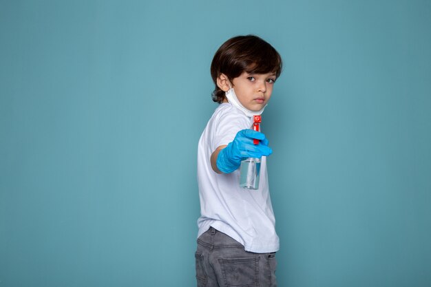Menino criança com spray nas mãos contra o coronavírus em luvas azuis na mesa azul
