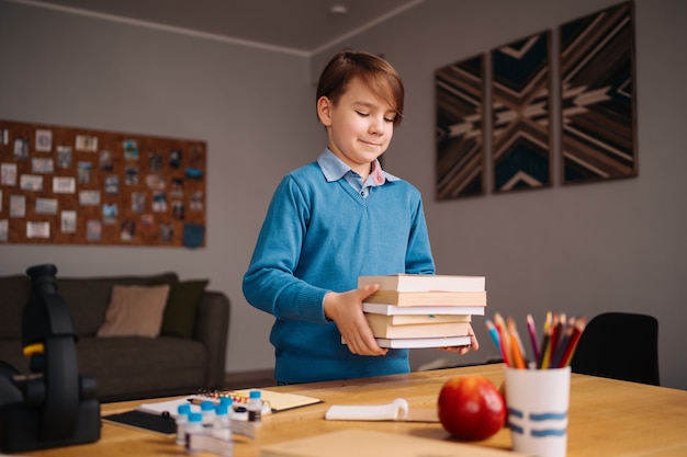 Menino da primeira série estudando em casa, segurando um monte de livros, se preparando para a aula online