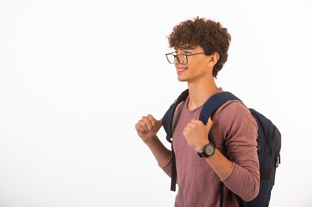 Menino de cabelo encaracolado em óculos de optique segurando sua mochila está feliz e alegre.