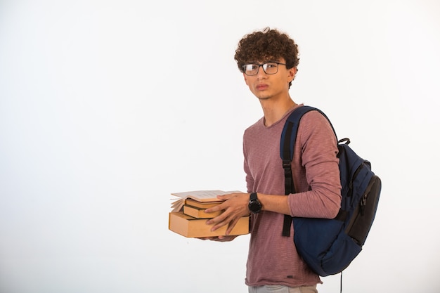 Menino de cabelo encaracolado em óculos opticos segurando livros escolares.