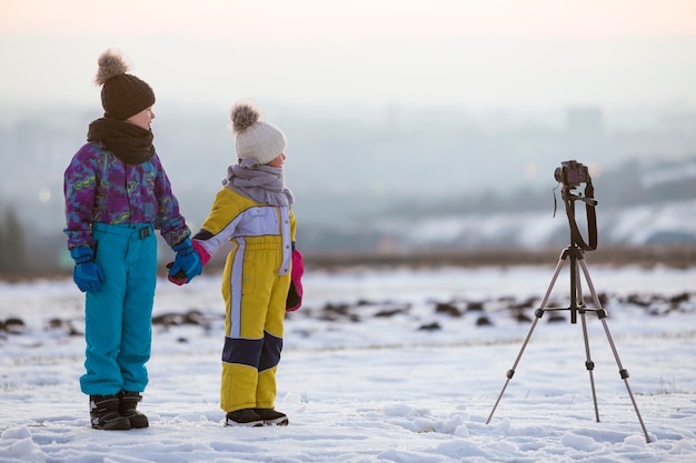 Menino de duas crianças e uma menina se divertindo do lado de fora no inverno, brincando com a câmera fotográfica em um tripé no campo coberto de neve.