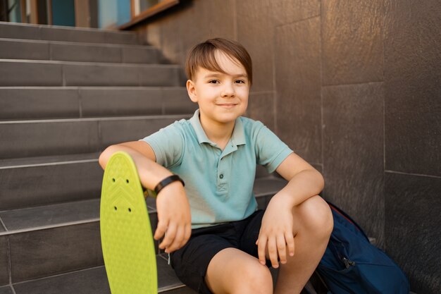 Menino de escola em camisa polo azul, sentado na escada com uma mochila azul e placa de centavo verde