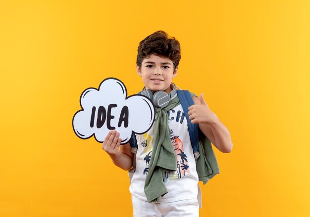 Menino de escola satisfeito com uma bolsa e fones de ouvido segurando uma bolha de ideia com o polegar isolado no fundo amarelo