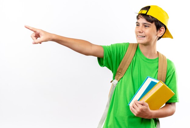 Menino de escola sorridente usando mochila com tampa segurando livros apontando ao lado