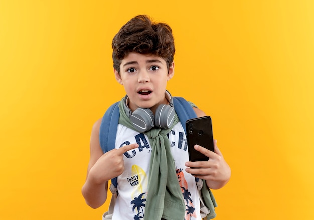 Menino de escola surpreso com uma bolsa nas costas e fones de ouvido segurando e apontando para o telefone