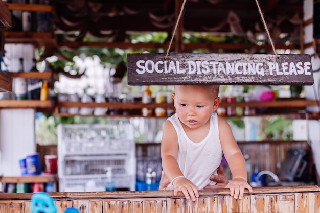 Menino de férias na tailândia com placa de distância social em bar