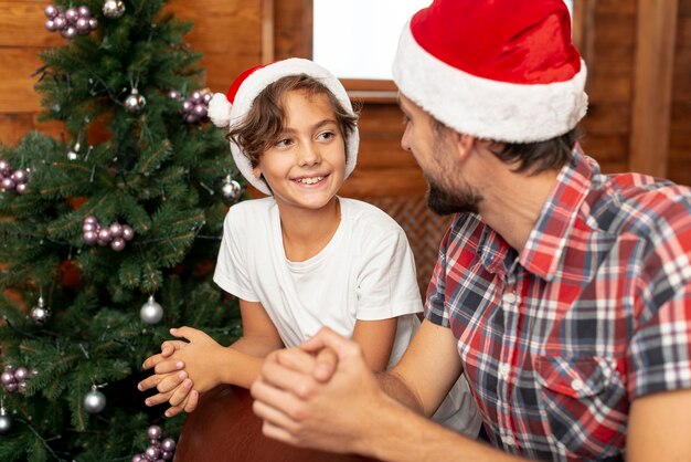 Menino de tiro médio com o pai sentado perto de árvore de Natal