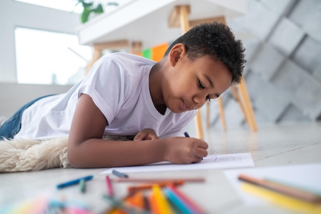 Menino deitado de bruços no chão desenhando com lápis