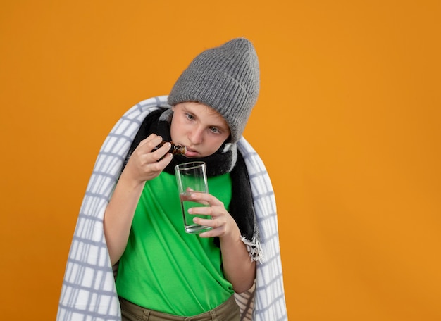 Menino doente com chapéu e lenço quente enrolado em um cobertor, pingando gotas de um frasco de remédio em um copo em cima da parede laranja