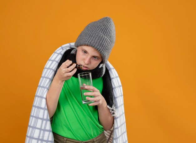 Menino doente com chapéu e lenço quente enrolado em um cobertor, pingando gotas de um frasco de remédio em um copo em cima da parede laranja