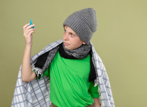 Menino doente com chapéu quente e lenço enrolado em cobertor segurando enemas olhando confuso e descontente em pé sobre uma parede de luz