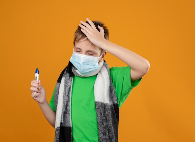 Menino doente com uma camiseta verde e um cachecol quente em volta do pescoço usando uma máscara protetora facial segurando um thremômetro parecendo confuso em pé sobre uma parede laranja