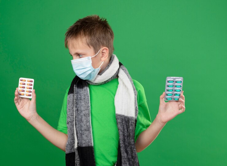 Foto grátis menino doente com uma camiseta verde e um lenço quente em volta do pescoço usando uma máscara protetora facial segurando comprimidos parecendo confuso tentando fazer a escolha em pé sobre um fundo verde