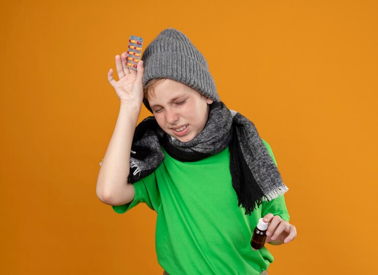 Foto grátis menino doente, vestindo uma camiseta verde com um lenço quente e um chapéu, sentindo-se mal, segurando um frasco de remédio e pílulas, sofrendo de dor de cabeça, doente e infeliz em pé sobre a parede laranja