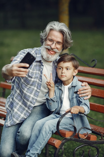 Foto grátis menino e avô sentado em um banco. família no parque. velho brincando com o neto. o avô usa um telefone.