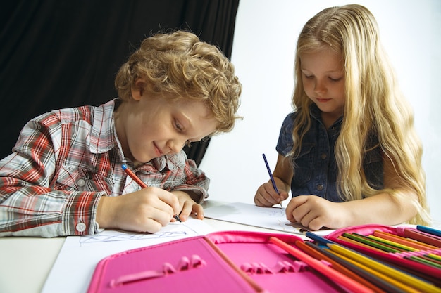 Menino e menina se preparando para a escola depois de uma longa pausa de verão. de volta à escola. pequenos modelos caucasianos desenhando juntos em fundo branco e preto. conceito de infância, educação, férias ou lição de casa.