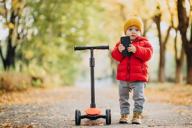 Menino em uma scooter segurando um telefone celular no parque outonal