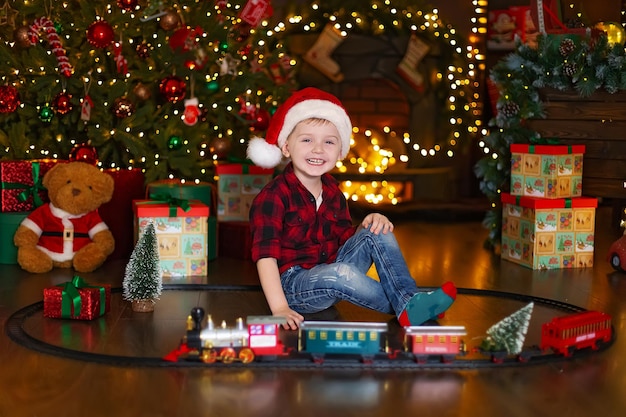 Menino feliz com chapéu de papai noel brincando com a locomotiva em casa enquanto está sentado perto da árvore de natal Foto Premium