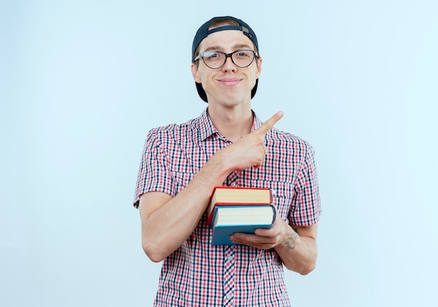 Menino feliz, jovem estudante, usando uma bolsa, óculos e boné, segurando livros e apontando para o lado em branco