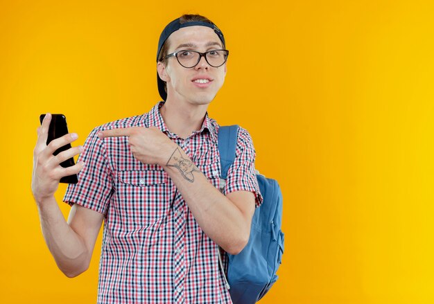 Menino feliz, jovem estudante vestindo uma bolsa, óculos e boné, segurando e apontando para o telefone em branco