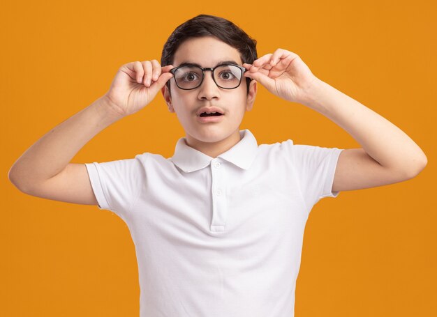 Menino impressionado usando e agarrando óculos, olhando diretamente isolado na parede laranja