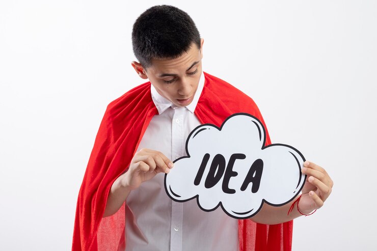 Foto grátis menino jovem super-herói impressionado com uma capa vermelha segurando e olhando para uma bolha de ideia isolada no fundo branco