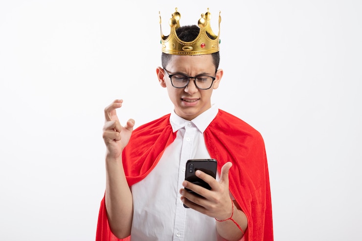 Foto grátis menino jovem super-herói irritado com capa vermelha usando óculos e uma coroa segurando e olhando para o telefone celular, mantendo a mão no ar isolado no fundo branco com espaço de cópia