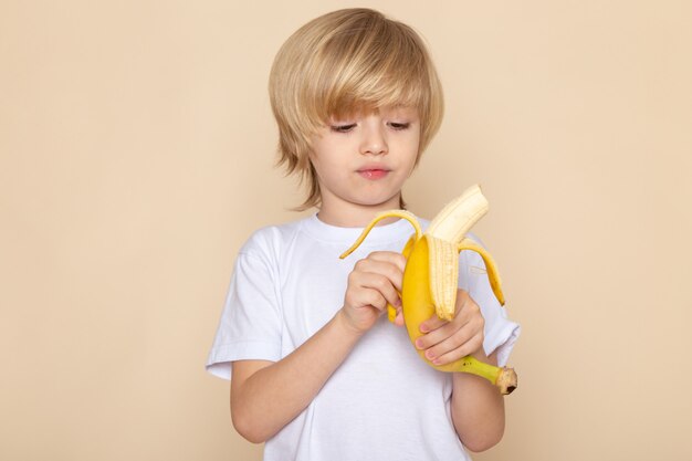 menino loiro bonito adorável descascar banana em camiseta branca na rosa