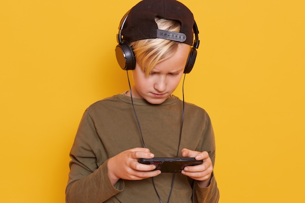 Menino loiro criança jogando jogos móveis no smartphone e usando a internet sem fio enquanto ouve música através de fones de ouvido