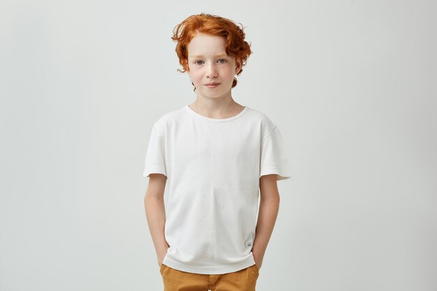 Menino ruivo bonito com um penteado bonito na camiseta branca, segurando as mãos nos bolsos, sorrindo gentilmente