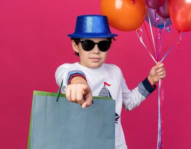 Menino satisfeito com chapéu de festa azul com óculos segurando balões e sacolas de presente, mostrando seu gesto isolado na parede rosa
