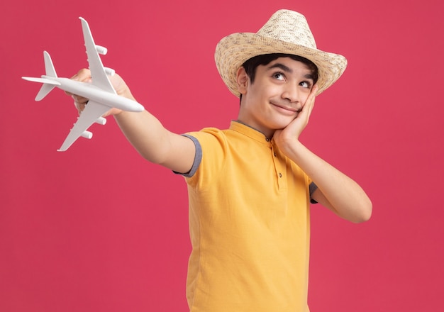 Menino satisfeito com chapéu de praia, estendendo-se para a frente do modelo do avião, colocando a mão no rosto, olhando para cima, isolado na parede rosa