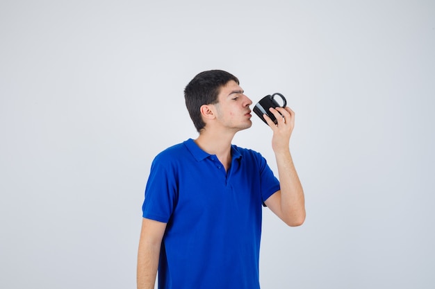 Menino segurando o copo, tentando beber o líquido com uma camiseta azul e parecendo sério