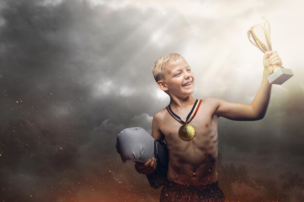 Menino sem camisa alegre segura luvas de boxer e a taça do vencedor fica no fundo de um céu nublado escuro que perfura os raios do sol.