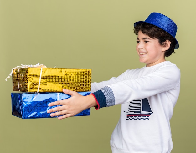 Menino sorridente com chapéu de festa azul segurando caixas de presente isoladas na parede verde oliva