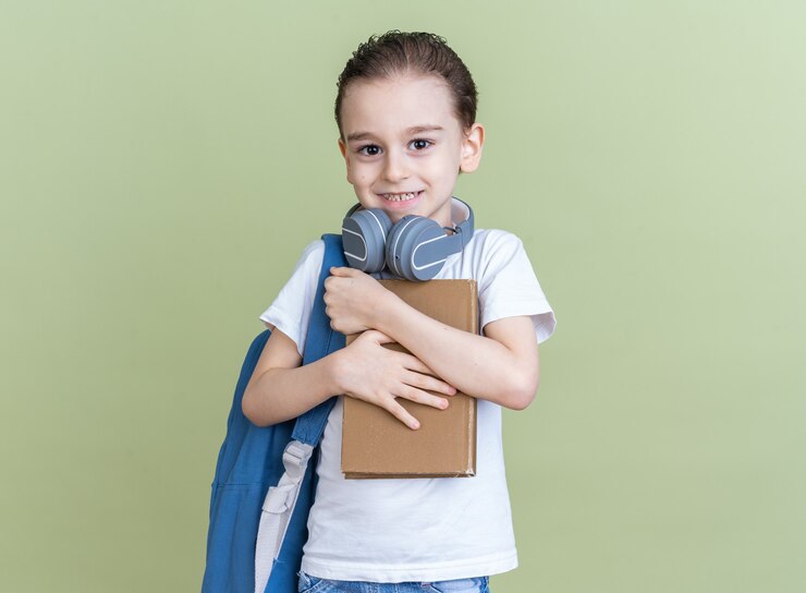 Foto grátis menino sorridente com mochila e fones de ouvido no pescoço segurando e abraçando o livro