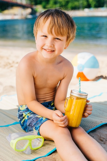 Menino sorridente com suco de vidro e sentado na praia