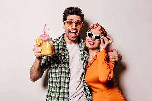 Menino travesso e menina em roupas elegantes e óculos escuros, abraçando, sorrindo e posando com um coquetel de laranja no espaço em branco.