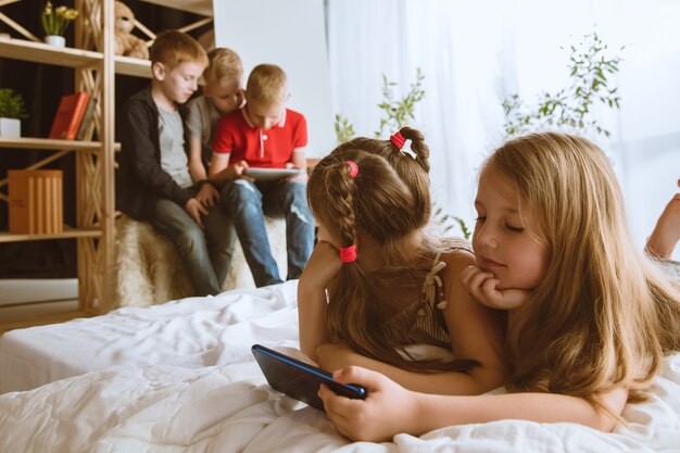 Meninos e meninas usando diferentes aparelhos em casa. Childs com relógios inteligentes, smartphone e fones de ouvido. Fazer selfie, bater papo, jogar, assistir vídeos. Interação de crianças e tecnologias modernas.