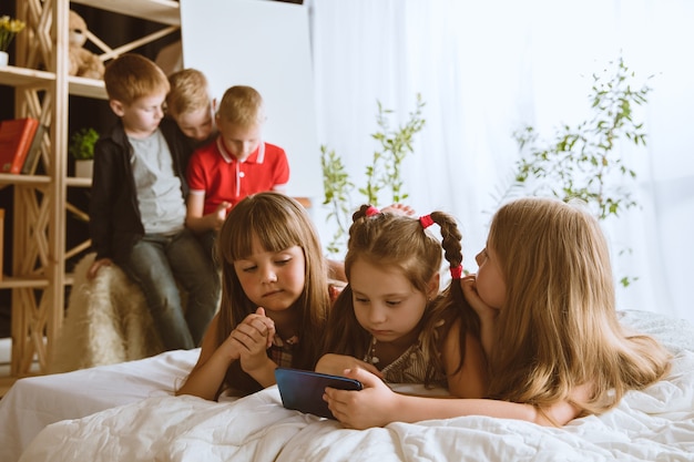 Meninos e meninas usando diferentes aparelhos em casa. Childs com relógios inteligentes, smartphone e fones de ouvido. Fazer selfie, bater papo, jogar, assistir vídeos. Interação de crianças e tecnologias modernas.