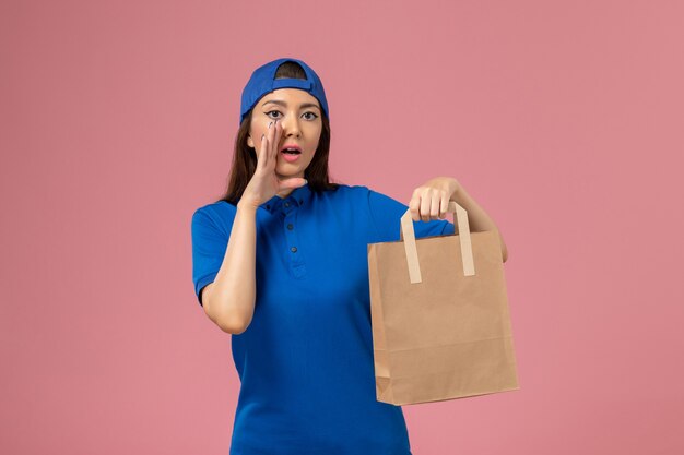 Mensageira de frente com capa uniforme azul segurando um pacote de entrega de papel chamando na parede rosa claro, prestadora de serviço prestando serviço