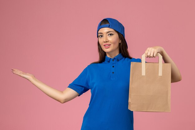 Mensageira de frente com capa uniforme azul segurando um pacote de papel na parede rosa claro, prestadora de serviço entregando o trabalho