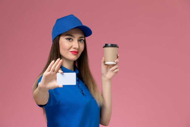 Mensageira de frente com uniforme azul e capa segurando a xícara de café de entrega e um cartão branco na parede rosa