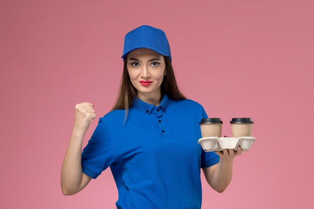 Mensageira de frente com uniforme azul e capa segurando xícaras de café comemorando na parede rosa