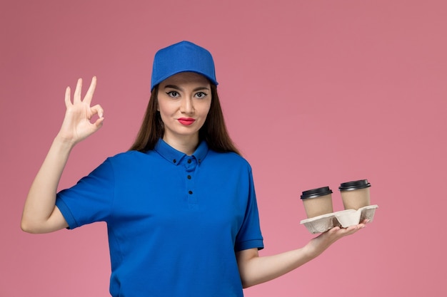 Mensageira de frente com uniforme azul e capa segurando xícaras de café marrom na mesa rosa