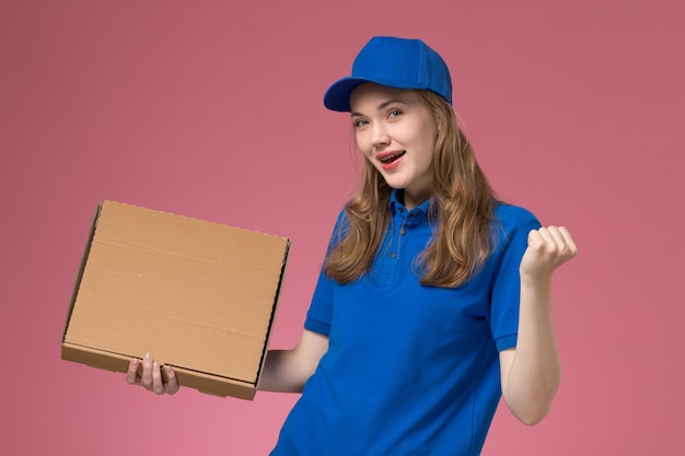 Mensageira de frente com uniforme azul segurando uma caixa de comida com expressão encantada na empresa de uniforme de serviço de trabalho de mesa rosa