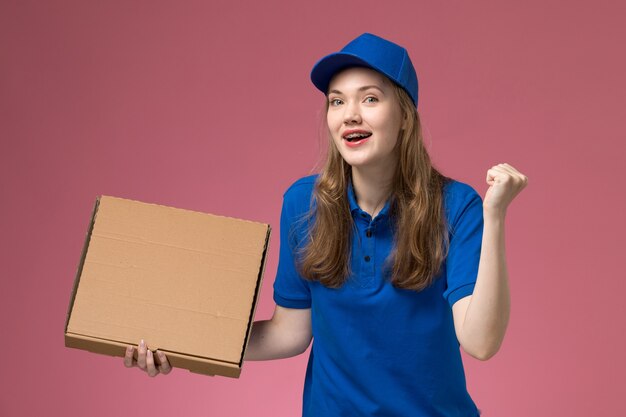 Mensageira de frente com uniforme azul segurando uma caixa de comida com expressão encantada na empresa de uniforme de serviço de trabalho de mesa rosa