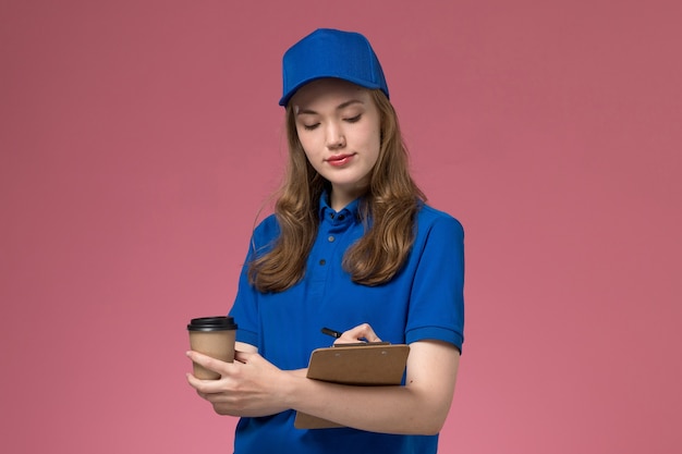 Foto grátis mensageira de frente com uniforme azul segurando uma xícara de café marrom escrevendo um bilhete no uniforme rosa claro da empresa de entrega de uniforme de serviço